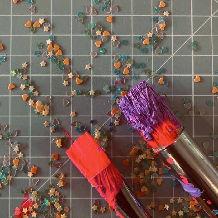 Two paint-loaded brushes resting on a cutting mat scattered with glitter stars and hearts