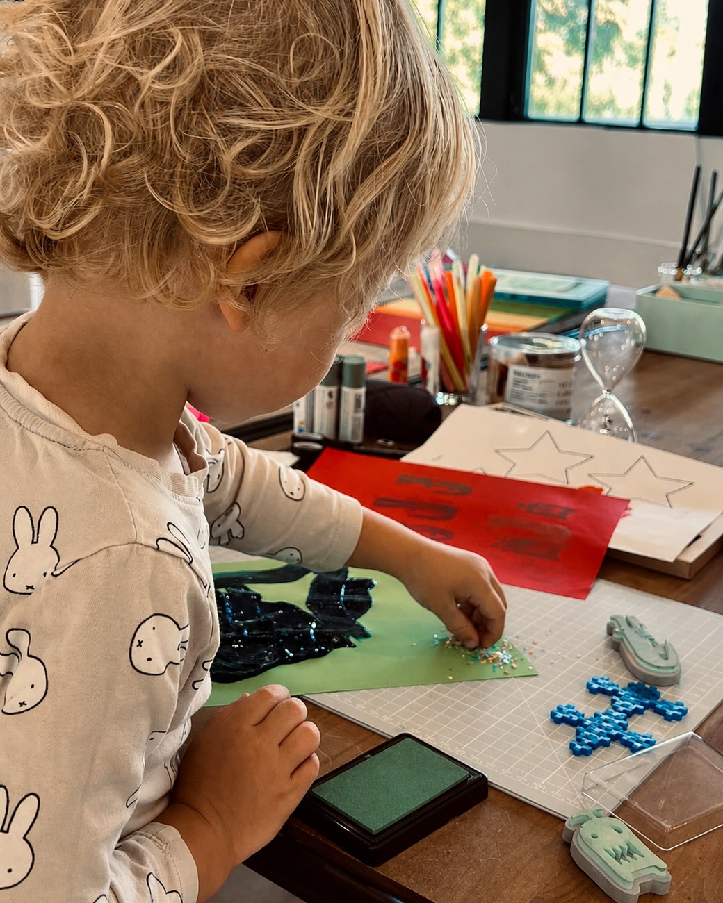 A young child, seen from behind, carefully sprinkling glitter onto a painted green page at the craft table