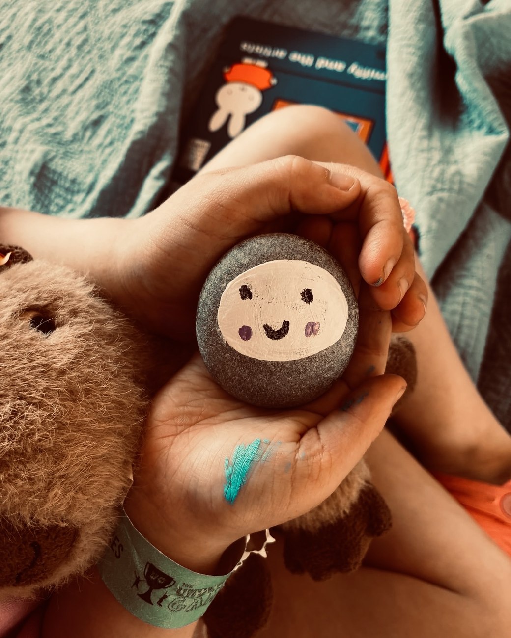 A child's cupped hands holding a pebble painted with a smiling face, a soft toy bear tucked beside them under a blanket