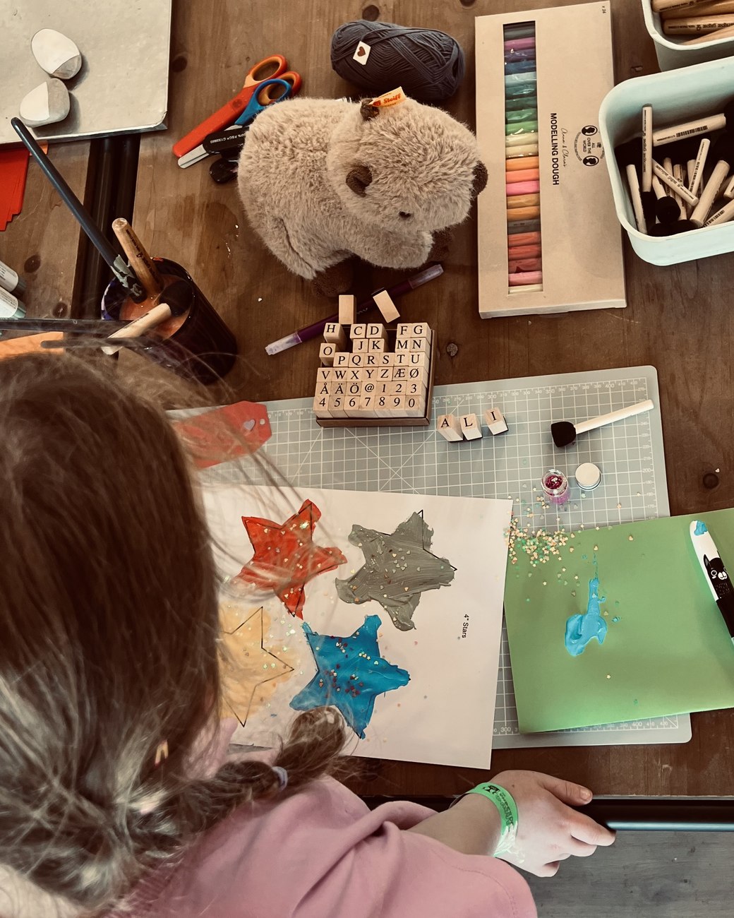 Seen from behind and above, a child at the craft table finishing a painting of three stars, surrounded by pastels, stamps and a soft toy