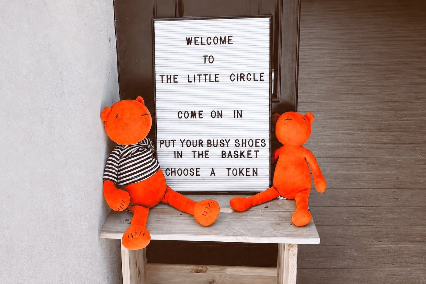 A letterboard sign reading: Welcome to The Little Circle, come on in, put your busy shoes in the basket, choose a token, with two soft toy bears sitting beside it
